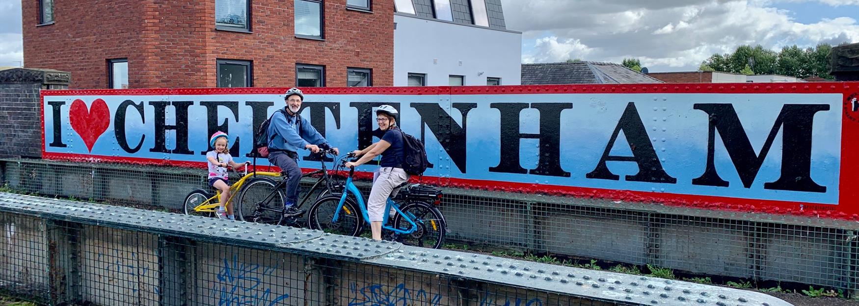 Family cycling along the Honeybourne Line in Cheltenham, with Cheltenham Paint Festival murals in background