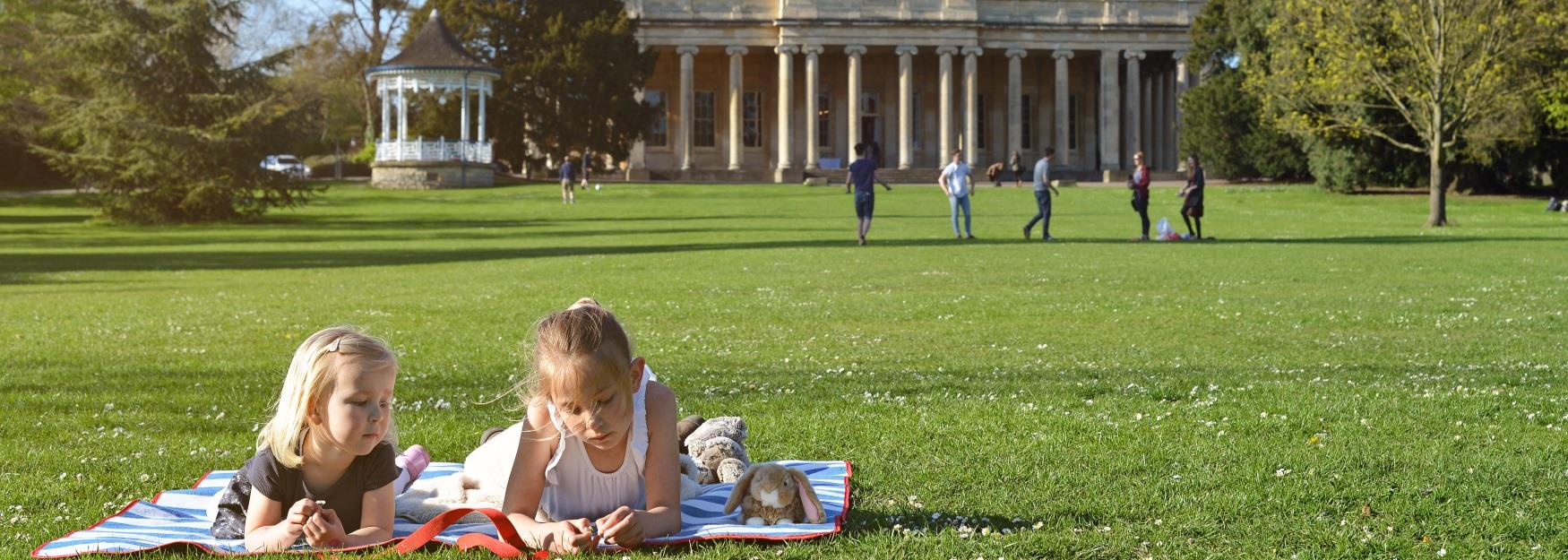 Two young children lying on a picnic blanket with Pittville Pump Room in the background